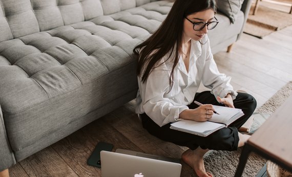 Wanita memakai baju putih sedang menulis sambil duduk bersila disamping laptop Macbook